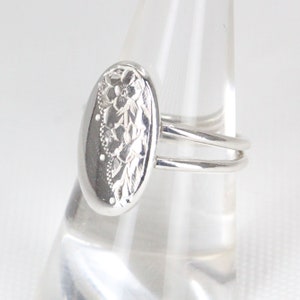 May include: A silver ring with a floral design on the top. The ring is oval shaped and has a double band.