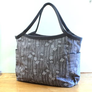 May include: A gray canvas tote bag with a white floral pattern. The bag has two wide straps and a large pocket on the side.