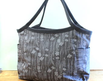 May include: A gray canvas tote bag with a white floral pattern. The bag has two wide straps and a large pocket on the side.