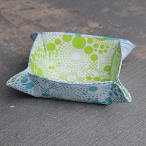 Puede incluir: Una pequeña cesta de almacenamiento cuadrada de tela con fondo blanco y un patrón de lunares verdes y azules. La cesta está doblada y tiene una ligera curva en los lados.