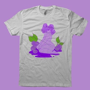 May include: White t-shirt with a purple illustration of a person and blueberries. The person has purple hair and is sitting in a pool of purple liquid, surrounded by green leaves and blueberries. The design is printed on a white t-shirt.