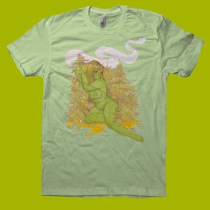 May include: Light green t-shirt featuring a green-skinned figure with long brown hair, surrounded by yellow-green plants and smoke. The illustration is detailed and artistic, with a unique style. The shirt is a solid light green color.