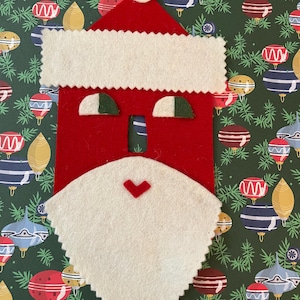 May include: A felt Santa Claus ornament with a red hat, white beard, and green eyes. The ornament is against a green background with a pattern of Christmas ornaments.