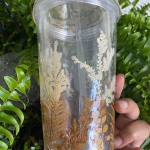 May include: A clear plastic tumbler with a straw, decorated with pressed dried ferns and leaves in shades of brown and gold.