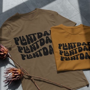 May include: Two brown t-shirts with the text "Plant Dad" in black, wavy letters. The shirts are laid out on a gray tiled surface.
