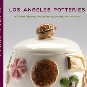 May include: A white ceramic cookie jar with a walnut-shaped lid. The jar is decorated with various brown, green, and pink designs, including cookies, flowers, and leaves. The text "LOS ANGELES POTTERIES" is printed on the front of the jar.