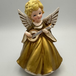 Può includere: Una statuetta di angelo in ceramica con capelli biondi, ali bianche e un abito color oro metallizzato. L'angelo tiene in mano un piccolo strumento a corde, un liuto, con entrambe le mani. La figura presenta dettagli del viso dipinti, inclusi occhi blu.