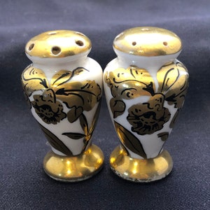 May include: A pair of white ceramic salt and pepper shakers with gold floral designs. The shakers have a round top with holes for dispensing salt and pepper.