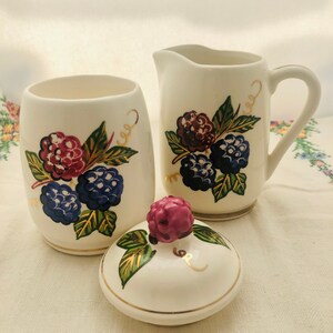 May include: A white ceramic sugar bowl and creamer set with a blackberry design. The set features gold accents and a pink blackberry on the lid of the sugar bowl.