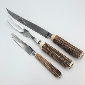 May include: A set of three silver-toned carving utensils with stag horn handles. The set includes a carving knife, a carving fork, and a smaller knife.