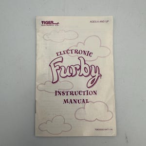 May include: A white instruction manual for an electronic Furby toy by Tiger Electronics, Ltd. The cover features purple text reading Electronic Furby Instruction Manual, the age rating Ages 6 and Up, and a pattern of light purple outlined clouds.