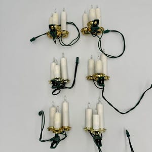 Peut inclure: Ensemble de six lumières de Noël en forme de bougie blanches avec des bases dorées et un fil vert. Chaque lumière a trois ampoules et un clip pour la fixer à un arbre ou à une autre surface.