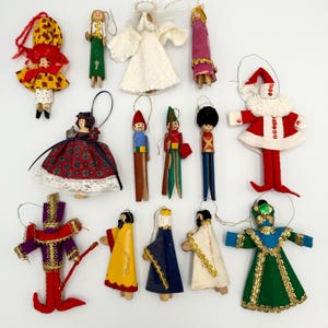 May include: A collection of 12 handmade Christmas ornaments featuring various characters, including a Santa Claus, an angel, a soldier, and a few figures in traditional clothing. The ornaments are made from felt, wood, and beads.