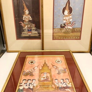Puede incluir: Tres pinturas enmarcadas que representan el arte tradicional tailandés. Las pinturas presentan figuras con vestimentas ornamentadas y tocados elaborados, sobre un fondo de oro y rojo.