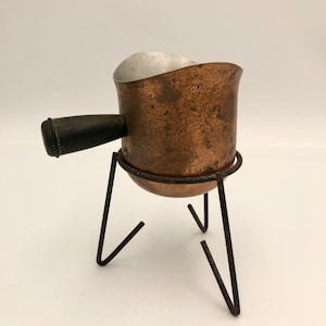 May include: A copper pot with a wooden handle sits on a black metal stand. The pot has a worn, aged look with a silver interior.