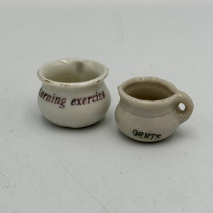 May include: Two small white ceramic chamber pots. One is labeled "Morning exercise" in purple lettering. The other is labeled "Gents" in black lettering.