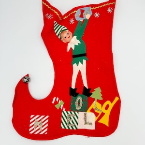 May include: Red felt Christmas stocking with a felt elf holding a gift. The elf is standing on a stack of felt blocks that spell "HOLIDAY".