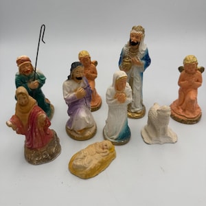 May include: A collection of nine small painted nativity figurines including a central infant in a manger, Mary, Joseph, a shepherd with a staff, a king, two kneeling angels, and a sheep, all featuring detailed facial expressions and colorful robes.
