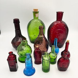 Puede incluir: Una colección de 12 botellas de vidrio vintage en varios colores, incluyendo rojo, verde y azul. Las botellas son de diferentes formas y tamaños, y algunas tienen diseños en relieve.