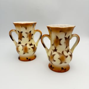 May include: Two small, white ceramic vases with brown floral designs and two handles. The vases are on a white background.
