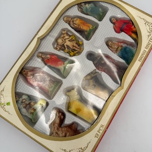 May include: A boxed set of 12 Shiny Brite nativity figurines featuring various characters including a baby in a manger, animals, and figures in colorful robes. The rectangular box has a clear plastic window and gold decorative border accents.