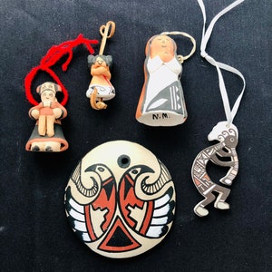 Puede incluir: Cinco adornos de cerámica hechos a mano con diseños nativos americanos. Los adornos representan a una mujer con un vestido blanco, un hombre sosteniendo a un niño, un pájaro con las alas extendidas, una figura de Kokopelli y una mujer con un tocado. Los adornos están pintados en negro, blanco, rojo y marrón.