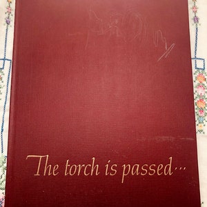 Puede incluir: Un libro granate con el texto dorado "The torch is passed...". El libro tiene una cubierta texturizada y parece ser de tapa dura. Hay marcas tenues en la parte superior del libro.
