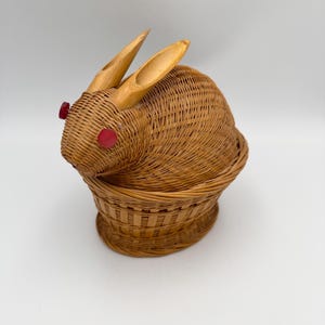 May include: A woven wicker basket shaped like a rabbit. The basket is brown with wooden ears and red circular eyes. The basket is designed to look like a rabbit sitting upright.