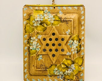 Puede incluir: Una rejilla de ventilación de pared de metal vintage con un diseño pintado de flores azules y hojas verdes. La rejilla tiene forma de estrella con un centro de malla y tiene un lazo para colgar en la parte superior. La rejilla está fechada "550 76".