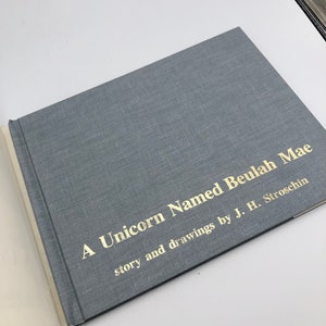 May include: A blue book with gold lettering on the cover. The title is "A Unicorn Named Beulah Mae" with the text "story and drawings by J. H. Stroschin" underneath.