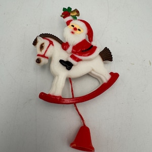 May include: A plastic Christmas ornament featuring Santa Claus riding a white rocking horse. Santa wears a red suit and hat, holding a small gold bell. The rocking horse has red rockers and a red bridle. A red string hangs down with a red bell.