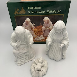 May include: A three-piece white porcelain nativity set featuring figures of a kneeling man with a staff, a kneeling woman, and a baby in a manger. The set is displayed in front of its original dark green box labeled Hand Crafted 3 Pcs Porcelain Nativity Set.