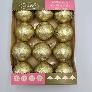 Op de afbeelding: Een doos met 12 gouden, glinsterende, onbreekbare kerstballen. De doos is gelabeld met "AMC" en "12 Glinsterende Onbreekbare Kerstballen".