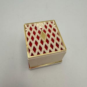 May include: A small, white box with a red and gold patterned lid. The lid has a diamond-shaped design with gold accents.