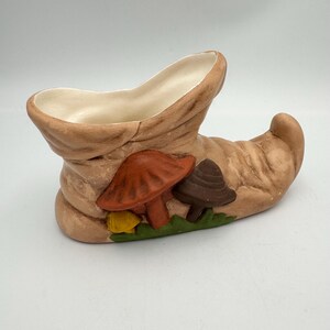 May include: A brown ceramic boot planter with three painted mushrooms. The mushrooms are red, brown, and yellow.