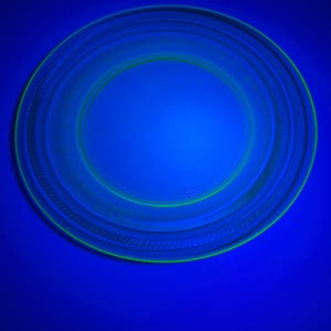 May include: A circular glass plate with a smooth centre and a textured, ribbed rim. Under ultraviolet light, the glass emits a vibrant, glowing neon green fluorescence against a deep blue background, highlighting the circular structure.