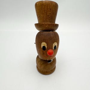 May include: A wooden figurine with a brown hat, brown face, black eyes, and a red nose.