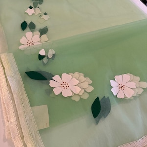 May include: A light green sheer fabric with white flowers and green leaves. The fabric has a white fringe border.