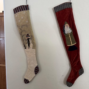 May include: Two decorative Christmas stockings hanging vertically. The left stocking is beige with a snowman and snowflake embroidery. The right stocking is deep red velvet featuring a Santa Claus figure. Both have striped cuffs, heels, and toes.