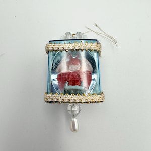 May include: A blue glass Christmas ornament with a white and gold trim. Inside the ornament is a red figurine of a person.