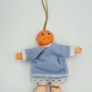 Pode incluir: Um pequeno ornamento de bebê de feltro azul com uma borda de renda branca e sapatos brancos. A boneca tem um rosto pintado com olhos castanhos e uma tez marrom clara. A boneca está pendurada em um cordão dourado.