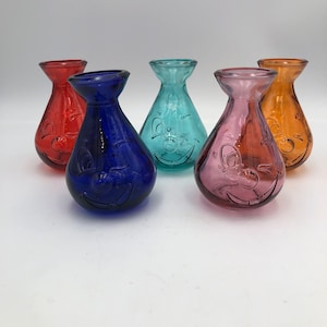 Puede incluir: Cinco jarrones de vidrio en diferentes colores, cada uno con una cara sonriente grabada en el vidrio. Los jarrones son rojo, azul, verde, morado y naranja.