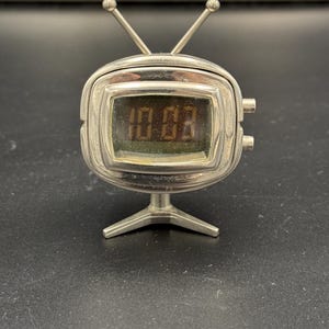 May include: A silver-toned digital clock shaped like a vintage television set with two antennas on top and a stand at the base. The screen displays the time 10:03 in dark digital numerals. Two small cylindrical buttons are located on the right side.