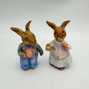 以下が含まれることがあります： セラミック製のウサギの置物2体。左側のウサギは青いズボンと灰色のジャケットを着て、ピンクの帽子を持っています。右側のウサギは白いドレスとピンクのエプロンを着て、果物の入ったバスケットを持っています。