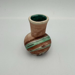 May include: A small, handcrafted ceramic vase with a bulbous base and a slender neck. The vase features a swirl pattern of brown, green, and white, with a green interior. The vase is likely used for decorative purposes.