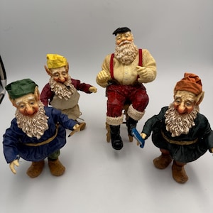 May include: A collection of four figurines featuring a central figure with a white beard, cream sweater, red suspenders, and red trousers, surrounded by three smaller gnome-like figures wearing colorful hats and tunics in blue, red, and dark green.