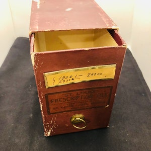 May include: A vintage brown wooden prescription file box with a brass lock and keyhole. The box has a label that reads "St. Louis Prescription File" and "Manufactured by St. Louis Label Works". The box also has a label with the numbers "1300 - 2400" and "23000".