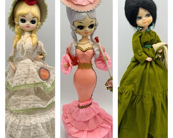 May include: Three vintage dolls are displayed, each in a unique gown. The first doll wears a white lace dress with green trim and a hat with a red rose. The second doll is in a pink dress with a matching hat and a parasol. The third doll is in a green dress.