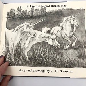 May include: A black and white illustration of three unicorns running through a field of grass. The text "A Unicorn Named Beulah Mae" is at the top of the image and the text "story and drawings by J. H. Stroschin" is at the bottom of the image.