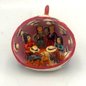May include: A ceramic ornament depicting a scene of four people sitting around a table with a dog. The ornament is painted in bright colors and has a pink interior. The people are wearing hats and the dog is brown and black.